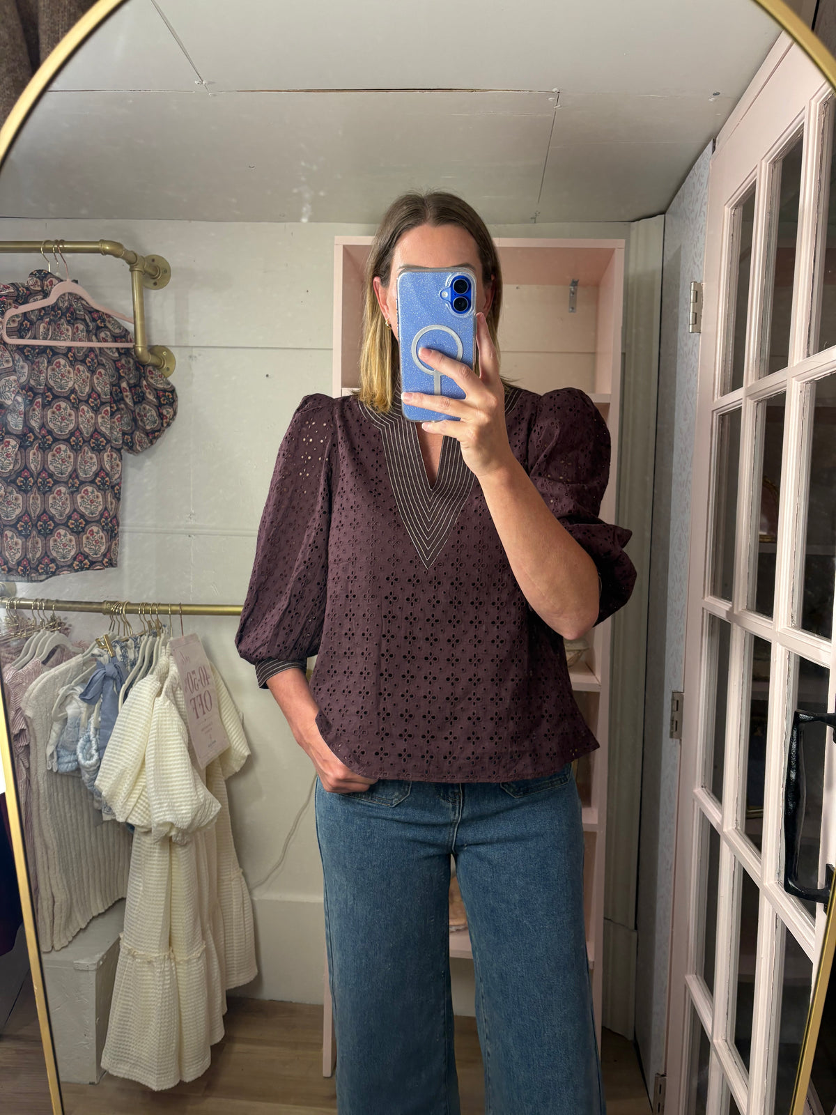 A woman takes a mirror selfie in a room showing a chocolate eyelet puff-sleeve blouse with a V-neck, paired with blue jeans.