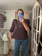 A woman takes a mirror selfie in a boutique showing a chocolate  eyelet blouse with puff sleeves and a V-neck, paired with blue jeans.