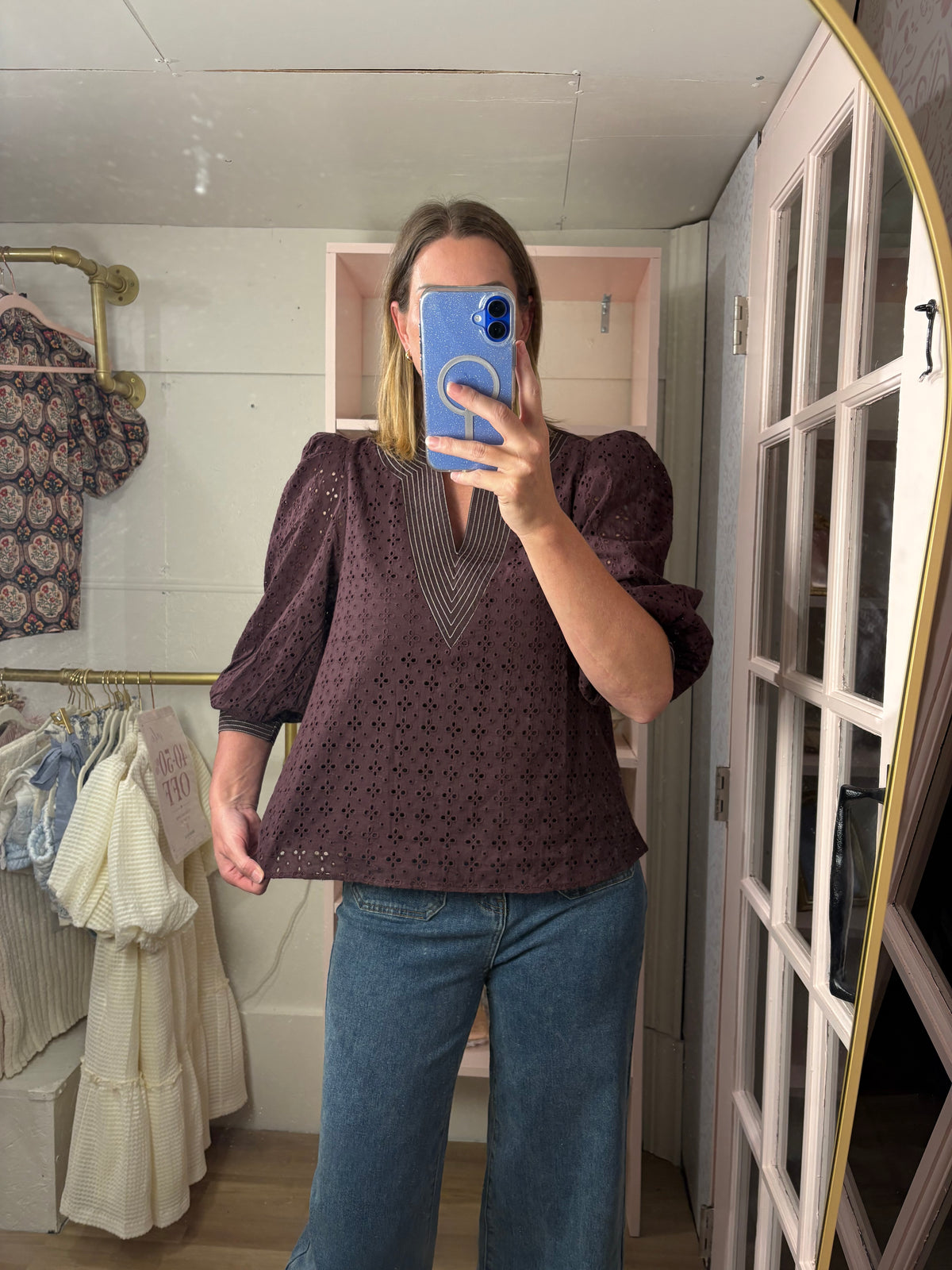 A woman takes a mirror selfie in a boutique showing a chocolate  eyelet blouse with puff sleeves and a V-neck, paired with blue jeans.