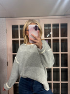 A woman takes a mirror selfie wearing a light gray knit sweater with white ribbed trim on the collar, cuffs, and hem, paired with dark blue jeans.