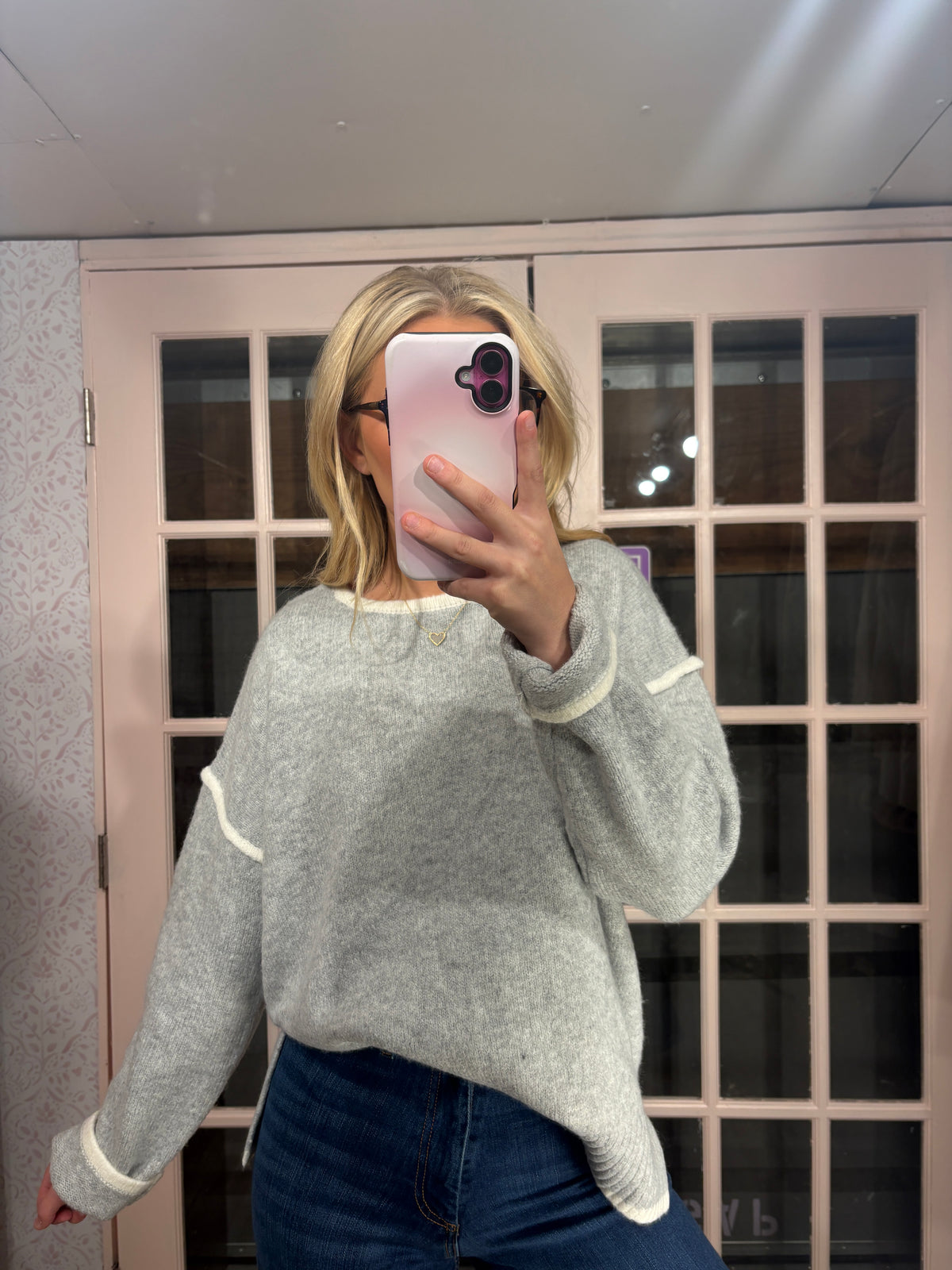 A woman takes a mirror selfie wearing a light gray knit sweater with white ribbed trim on the collar, cuffs, and hem, paired with dark blue jeans.