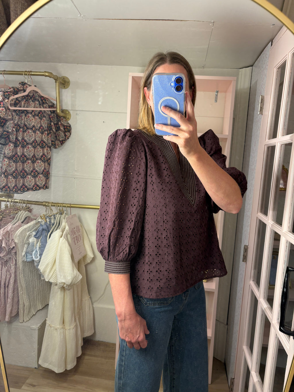 A woman taking a mirror selfie in a boutique, wearing a chocolate  eyelet puff-sleeve blouse and blue jeans.