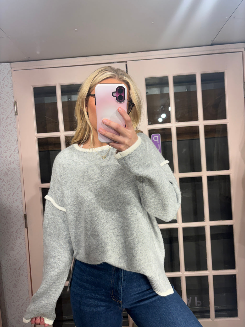 A woman takes a mirror selfie wearing a light gray fuzzy knit sweater with white ribbed trim and blue jeans, standing in front of a pink-framed glass door.