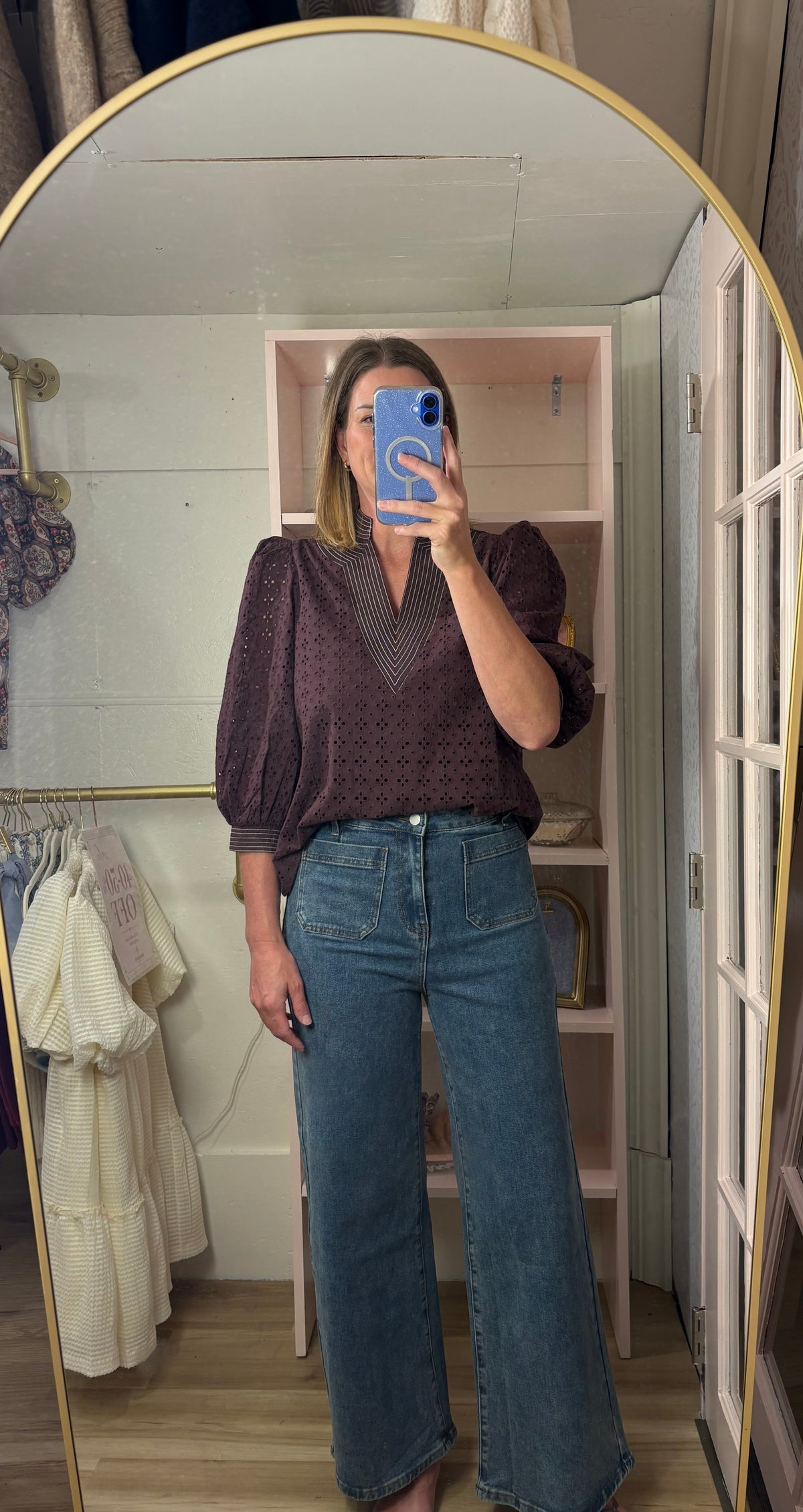 A woman takes a mirror selfie in a fitting room wearing a dark purple patterned puff-sleeve blouse and high-waisted wide-leg blue jeans.