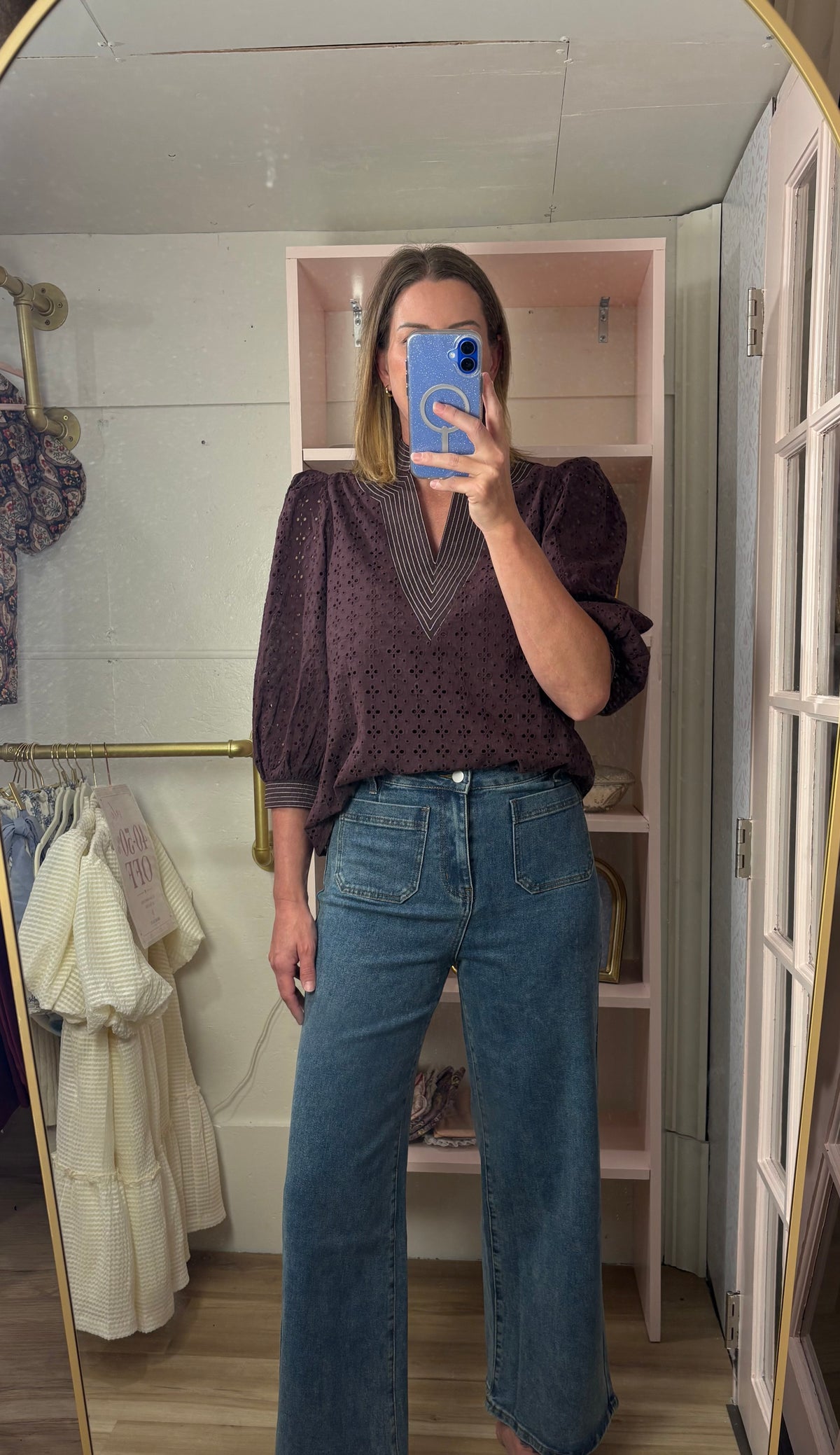 A woman takes a mirror selfie in a fitting room wearing a dark purple textured puff-sleeve blouse and high-waisted blue wide-leg jeans.