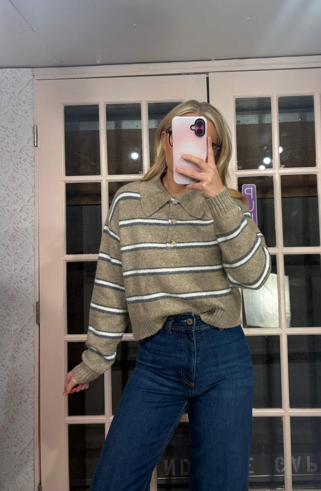 A woman takes a mirror selfie wearing a beige and white striped knit polo sweater with blue jeans, standing in front of a pink-framed glass door.