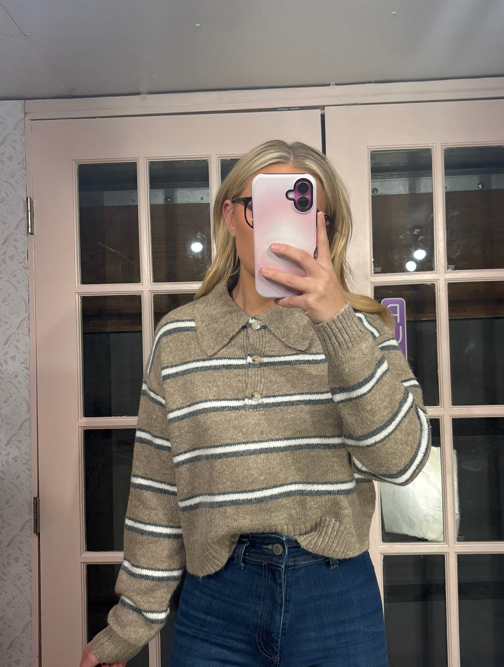 A woman takes a mirror selfie wearing a beige knit polo sweater with thin white horizontal stripes and blue jeans, standing in front of a pink-framed glass door.