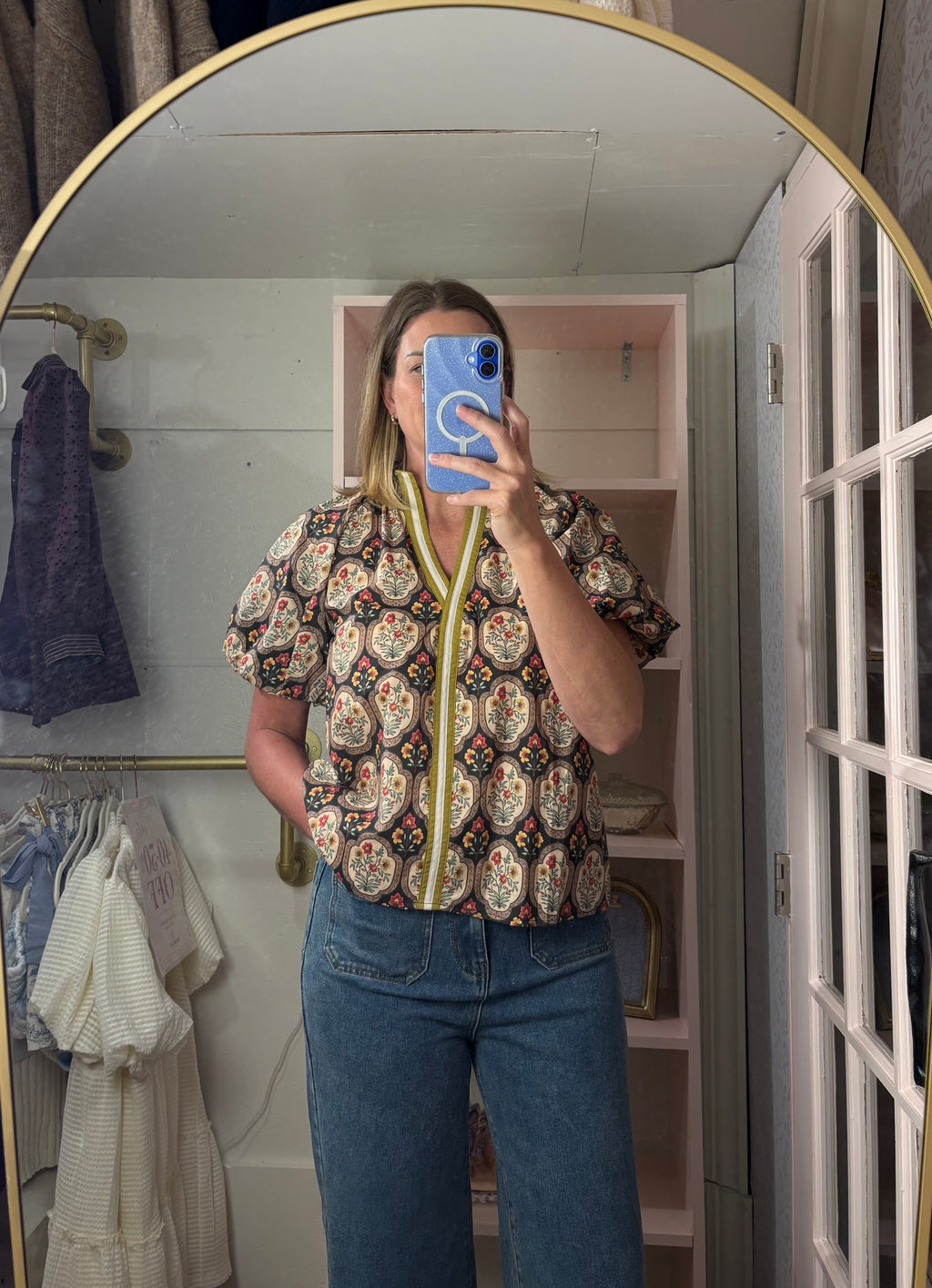 A woman takes a mirror selfie in a room, wearing a short-sleeve button-up blouse with a colorful paisley pattern and a lime green split neckline, paired with blue jeans.