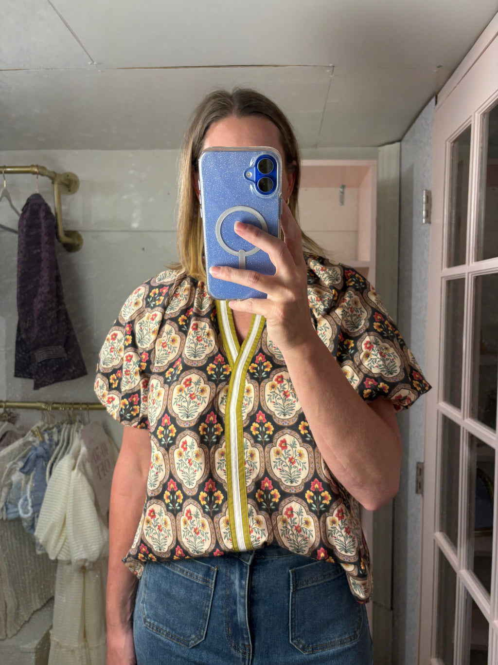 A woman taking a mirror selfie in a boutique, wearing a short-sleeve button-up blouse with a colorful paisley pattern and a yellow zipper detail, paired with blue jeans.