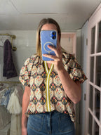 A woman taking a mirror selfie in a boutique, wearing a short-sleeve button-up blouse with a colorful paisley pattern and a yellow zipper detail, paired with blue jeans.