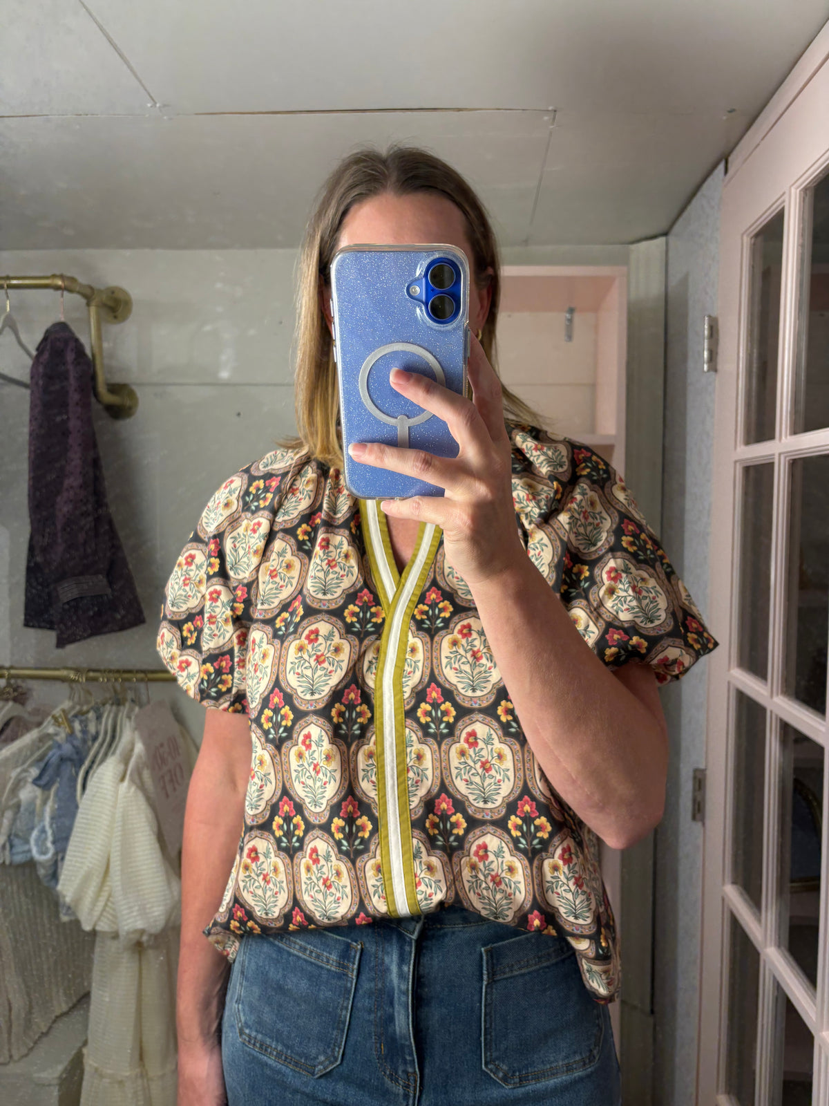 A woman taking a mirror selfie in a boutique, wearing a short-sleeve button-up blouse with a colorful paisley pattern and a yellow zipper detail, paired with blue jeans.