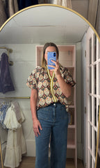 A woman takes a mirror selfie in a fitting room wearing a short-sleeve button-up shirt with a colorful paisley pattern and blue jeans.