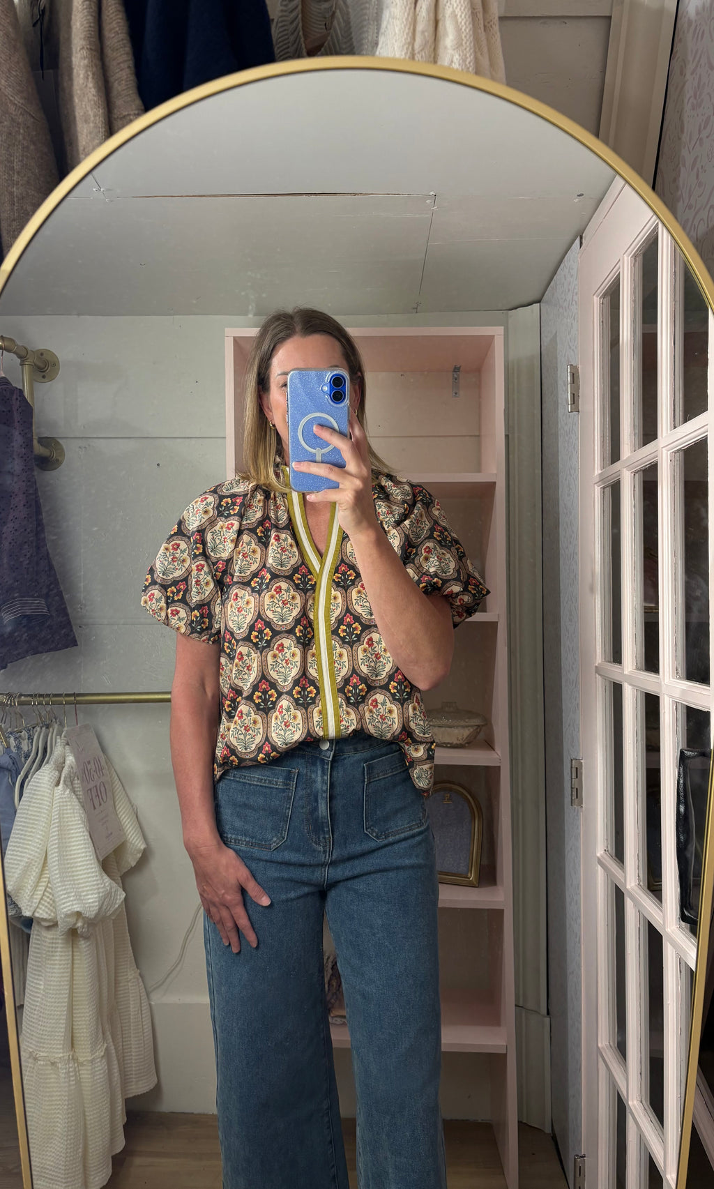 A woman takes a mirror selfie in a dressing room wearing a short-sleeve button-up shirt with a colorful paisley pattern and blue jeans.