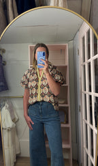 A woman takes a mirror selfie in a room, wearing a short-sleeve button-up shirt with a colorful paisley pattern and blue wide-leg jeans. The shirt features a yellow zipper detail at the collar.