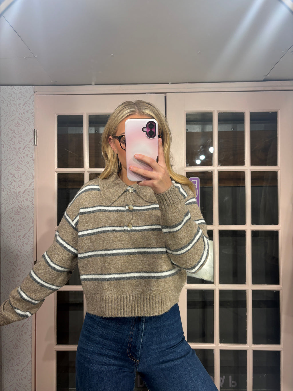 A woman takes a mirror selfie wearing a beige knit sweater with thin white and dark gray horizontal stripes, paired with blue jeans.