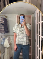 A woman takes a mirror selfie in a boutique while wearing a short-sleeve plaid button-up blouse with puff sleeves and blue jeans. The boutique has a pink shelving unit and clothing racks in the background.