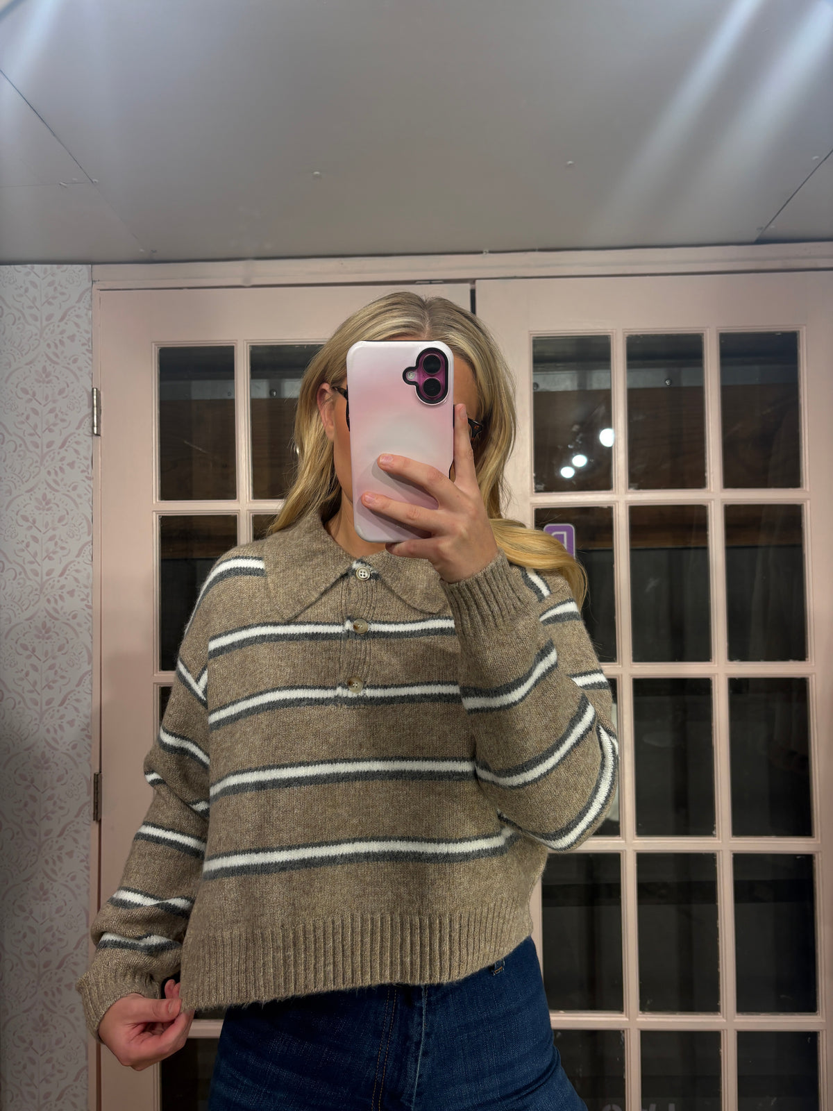 A woman takes a mirror selfie wearing a beige knit sweater with thin white and dark gray horizontal stripes, paired with blue jeans. The background shows a light-colored paneled door.