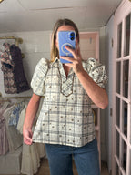 A woman takes a mirror selfie in a boutique wearing a cream-colored short-sleeve blouse with a black and gray plaid pattern, brown buttons, and puff sleeves, paired with blue jeans.