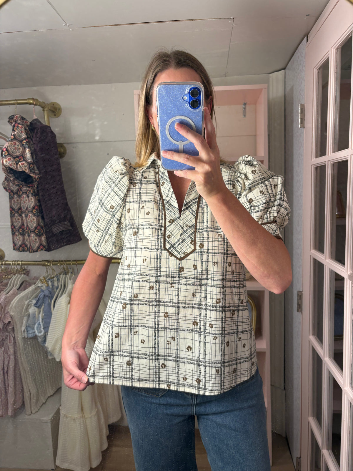 A woman takes a mirror selfie in a boutique wearing a cream-colored short-sleeve blouse with a black and gray plaid pattern, brown buttons, and puff sleeves, paired with blue jeans.
