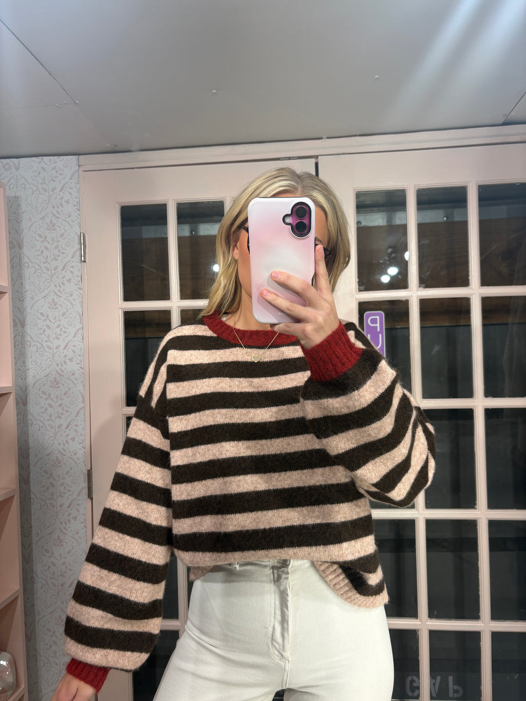 A woman takes a mirror selfie wearing a beige and dark brown horizontally striped fuzzy knit sweater with red ribbed collar and cuffs, paired with white pants.