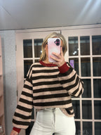 A woman takes a mirror selfie wearing a beige and dark brown horizontally striped fuzzy knit sweater with red ribbed collar and cuffs, paired with white pants.