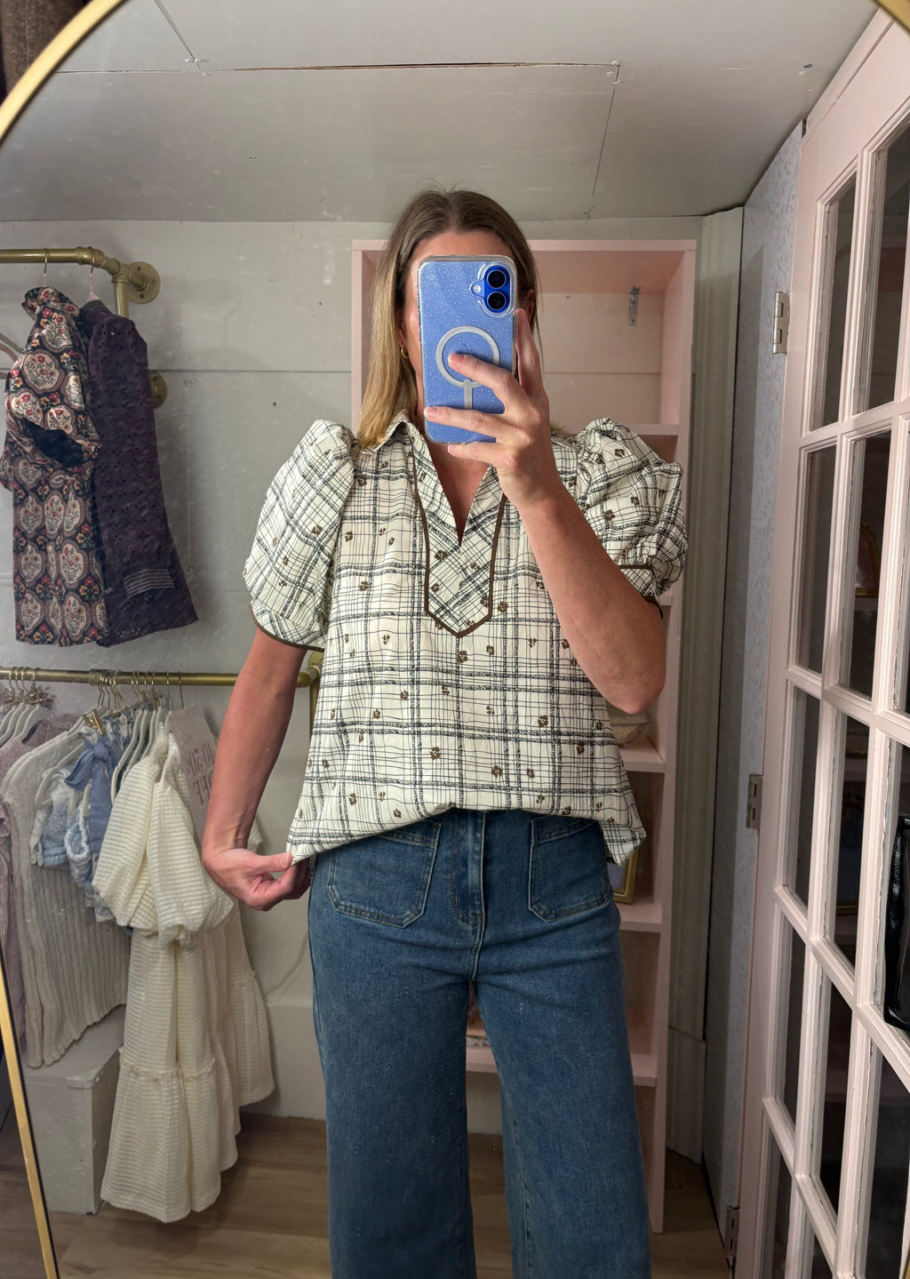A woman takes a mirror selfie in a boutique wearing a cream and black plaid short-sleeve button-up blouse with puff sleeves and blue jeans.