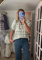 A woman takes a mirror selfie in a boutique wearing a cream and black plaid short-sleeve button-up blouse with puff sleeves and blue jeans.