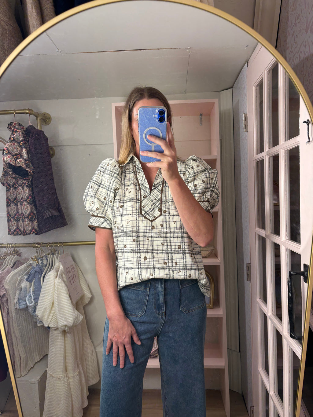A woman takes a mirror selfie in a boutique showing a short-sleeve plaid button-up shirt with puff sleeves and blue jeans.