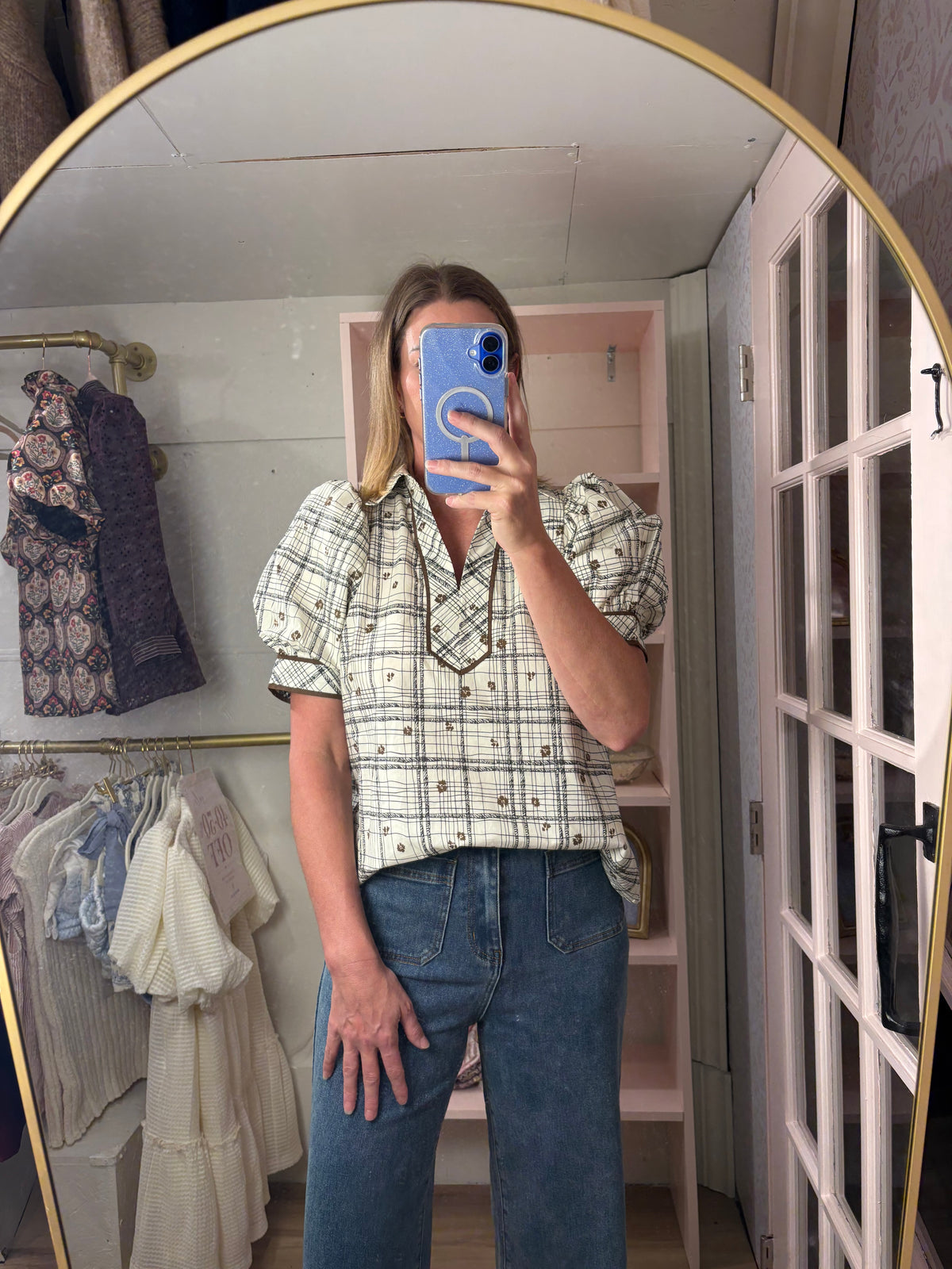 A woman takes a mirror selfie in a boutique showing a short-sleeve plaid button-up shirt with puff sleeves and blue jeans.