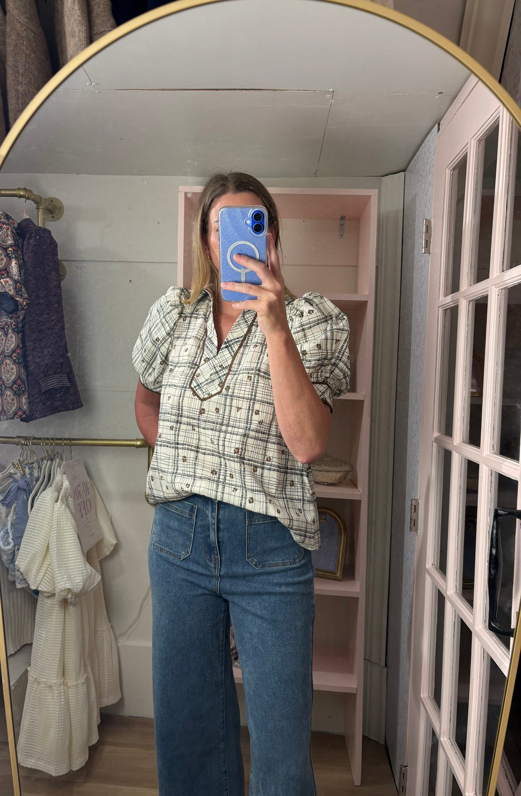 A woman taking a mirror selfie in a room, wearing a short-sleeve plaid button-up shirt in beige, white, and black, paired with blue jeans.