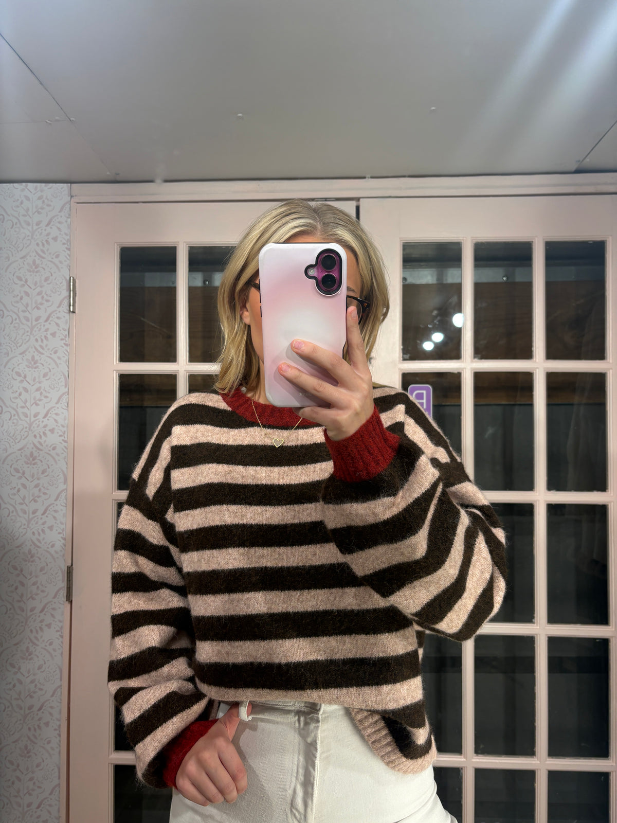 A person taking a mirror selfie wearing a fuzzy knit sweater with horizontal black and beige stripes, featuring a contrasting red crew neck and cuffs, paired with white pants.