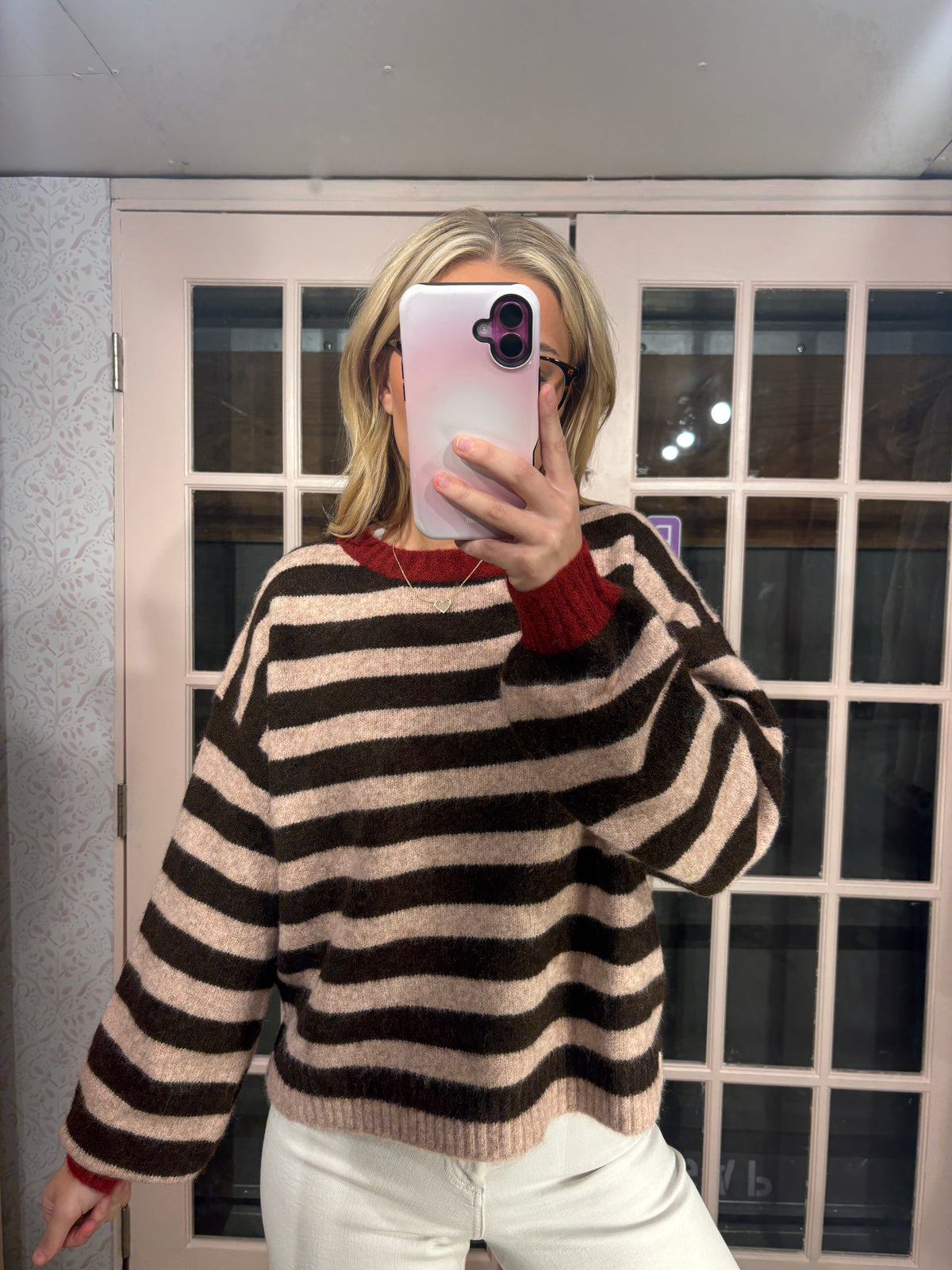 A woman takes a mirror selfie wearing a brown and beige horizontally striped knit sweater with a red crewneck undershirt and white pants, standing in front of a white-framed glass door.