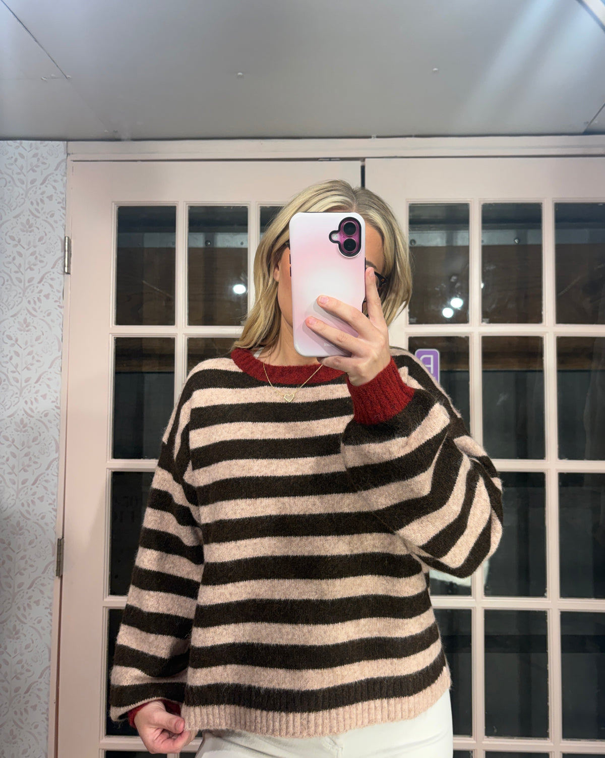 A woman takes a mirror selfie wearing a fuzzy knit sweater with horizontal black and beige stripes, featuring a contrasting dark red crew neck collar and cuffs. She is standing in front of a white-framed glass door.