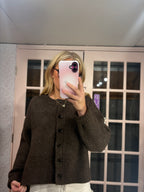 A woman takes a mirror selfie wearing a dark brown knit button-up cardigan sweater and white pants, standing in front of a pink-framed glass door.