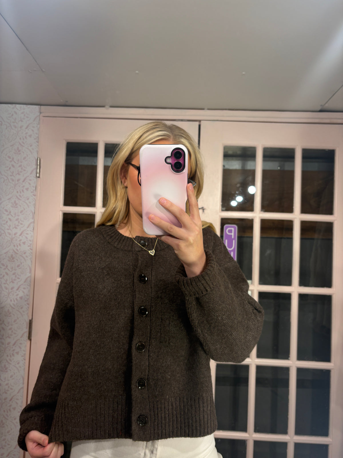 A woman takes a mirror selfie wearing a dark brown knit button-up cardigan sweater and white pants, standing in front of a pink-framed glass door.
