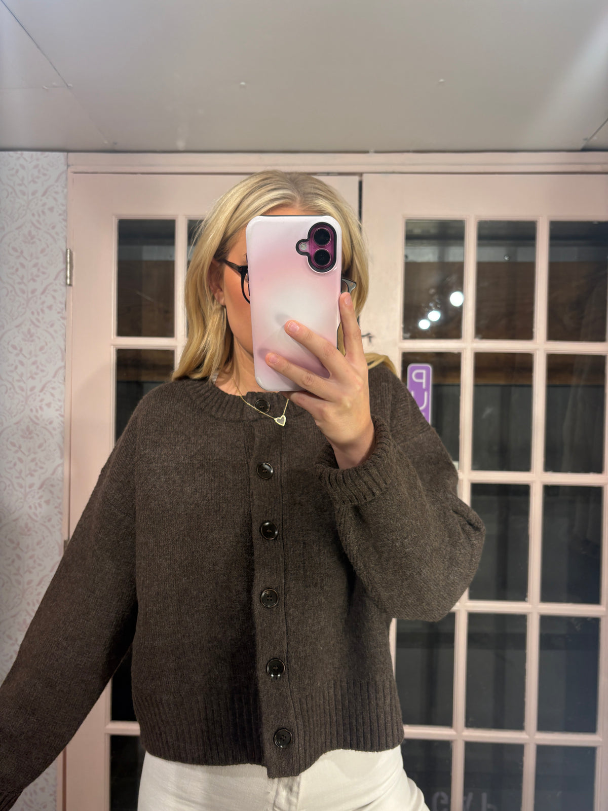 A woman takes a mirror selfie wearing a dark brown button-up knit cardigan sweater and white pants. She is holding a pink phone in front of her face, with a light-colored paneled door in the background.