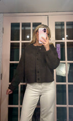 A woman takes a mirror selfie wearing a dark brown cropped button-up cardigan sweater and white pants in front of a glass-paneled door.