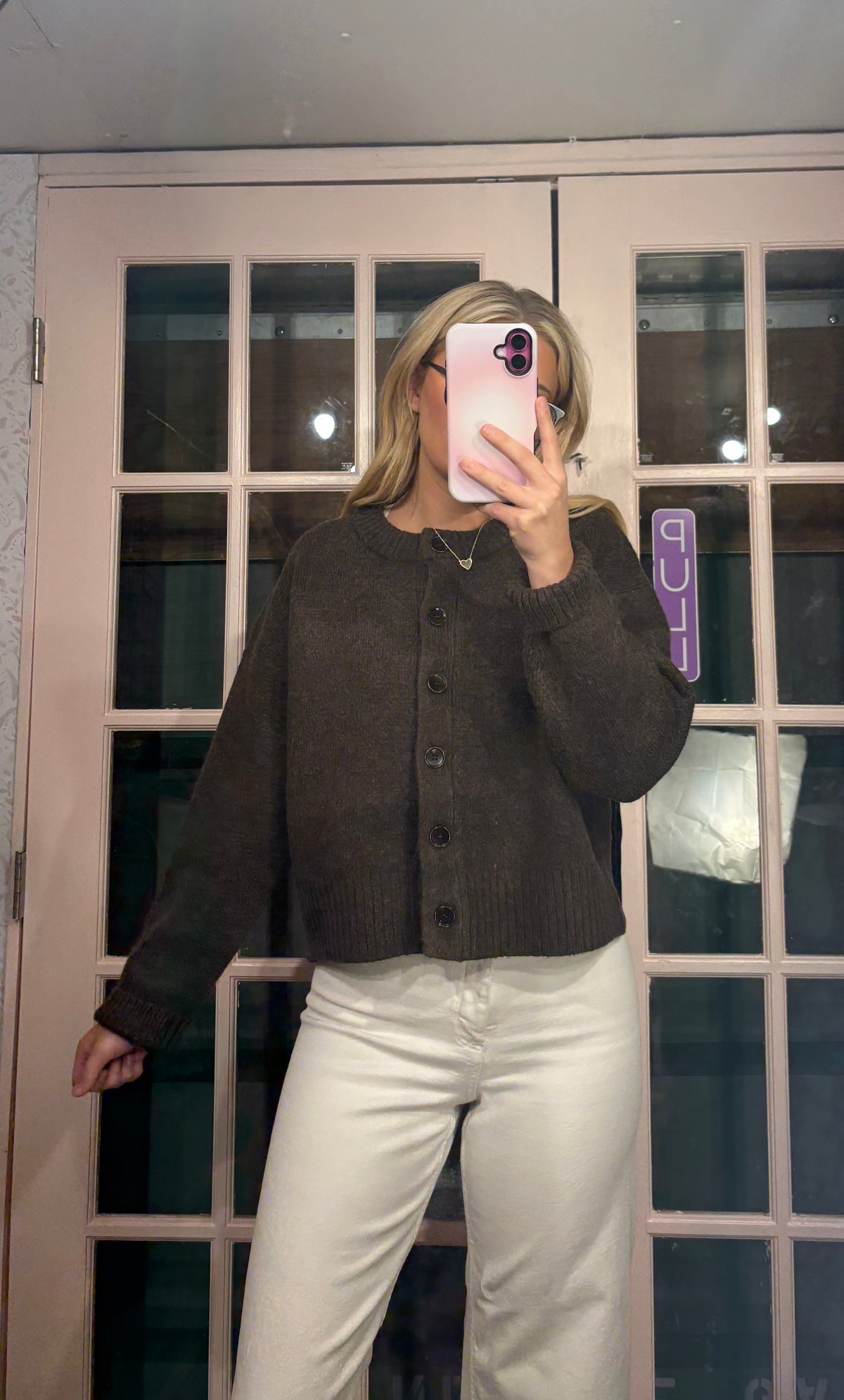 A woman takes a mirror selfie wearing a dark brown cropped button-up cardigan sweater and white pants in front of a glass-paneled door.