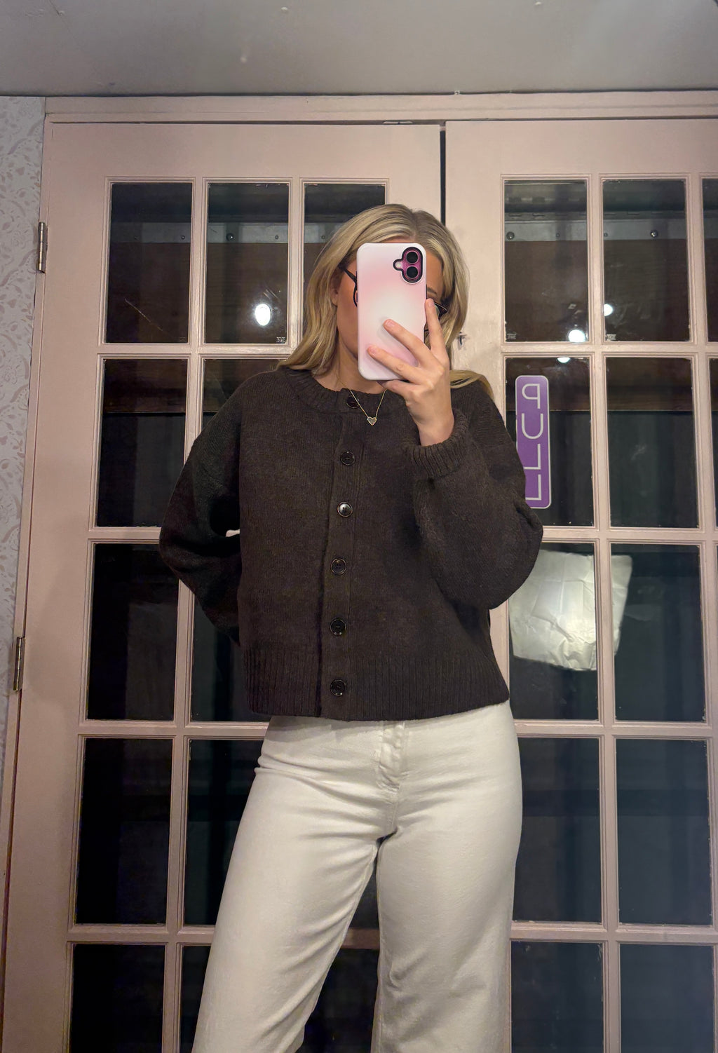 A woman takes a mirror selfie wearing a dark brown button-up knit cardigan and white pants in front of a pink-framed glass door.