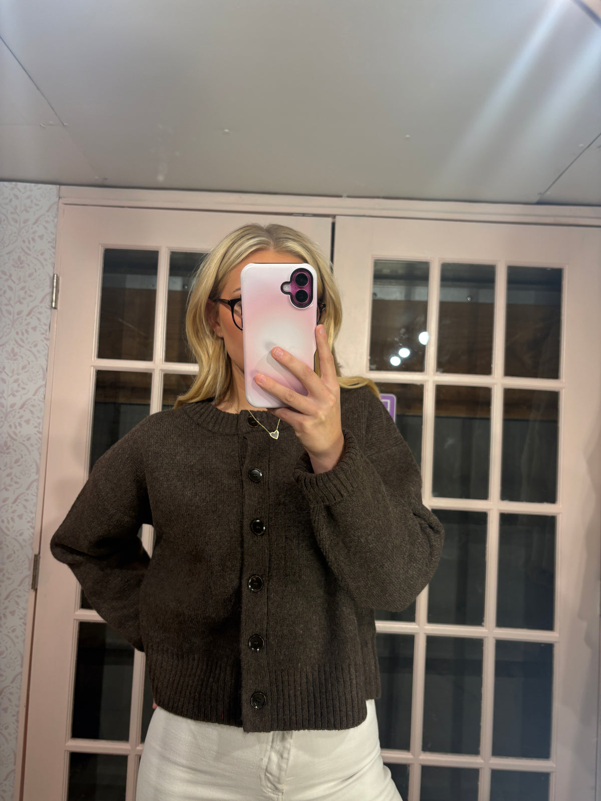 A woman takes a mirror selfie wearing a dark brown knit button-up cardigan sweater and white pants, standing in front of a pink glass-paneled door.