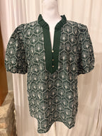 A dark green short-sleeve blouse with a white geometric and floral pattern, featuring a ruffled mock neck and button placket, displayed on a mannequin.