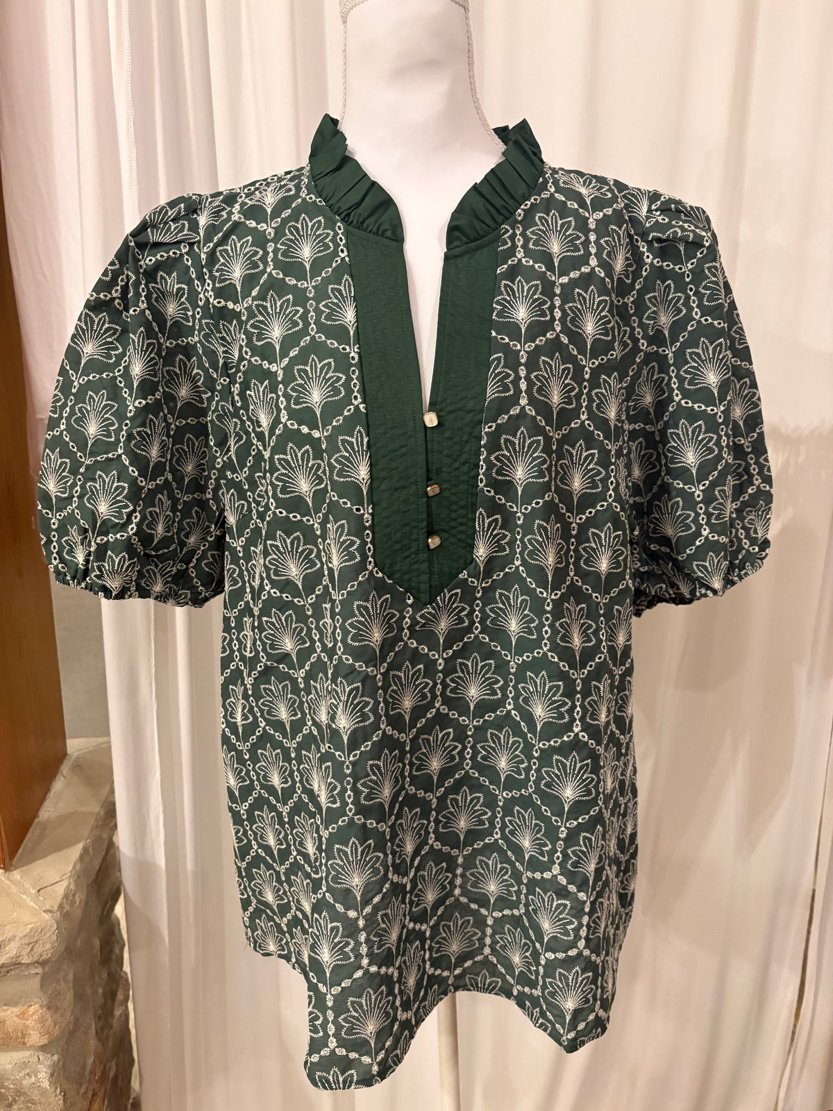 A dark green short-sleeve blouse with a white geometric and floral pattern, featuring a ruffled mock neck and button placket, displayed on a mannequin.