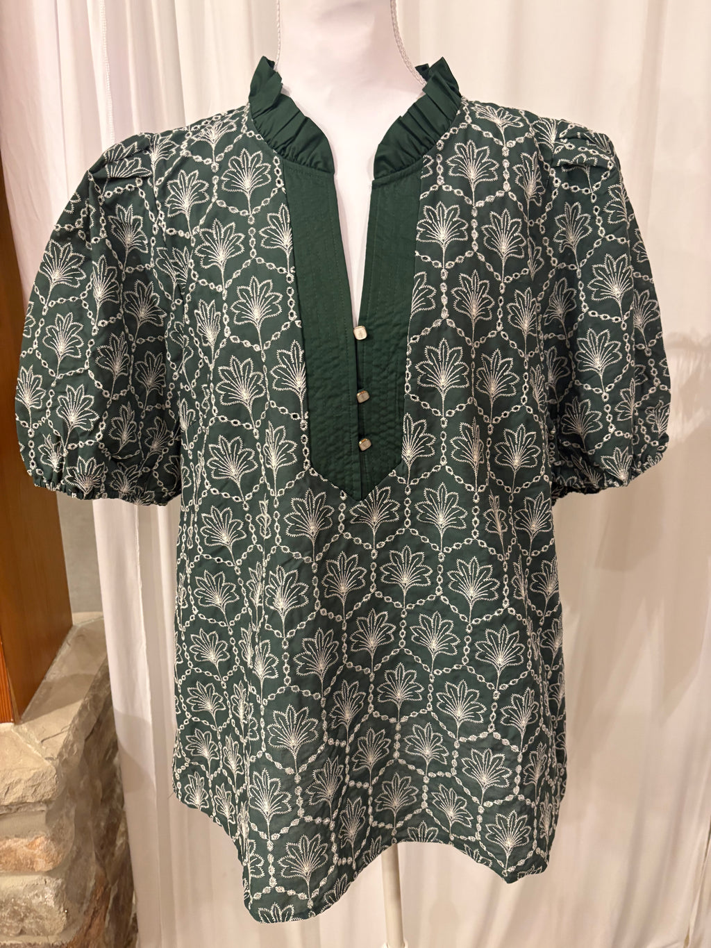 A dark green short-sleeve blouse with a white geometric and floral pattern, displayed on a mannequin torso. The blouse features a ruffled mock neck, a buttoned placket with light-colored buttons, and puffed short sleeves.