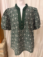 A dark green short-sleeve blouse with a white geometric and floral pattern, displayed on a mannequin torso. The blouse features a ruffled mock neck, a buttoned placket with light-colored buttons, and puffed short sleeves.