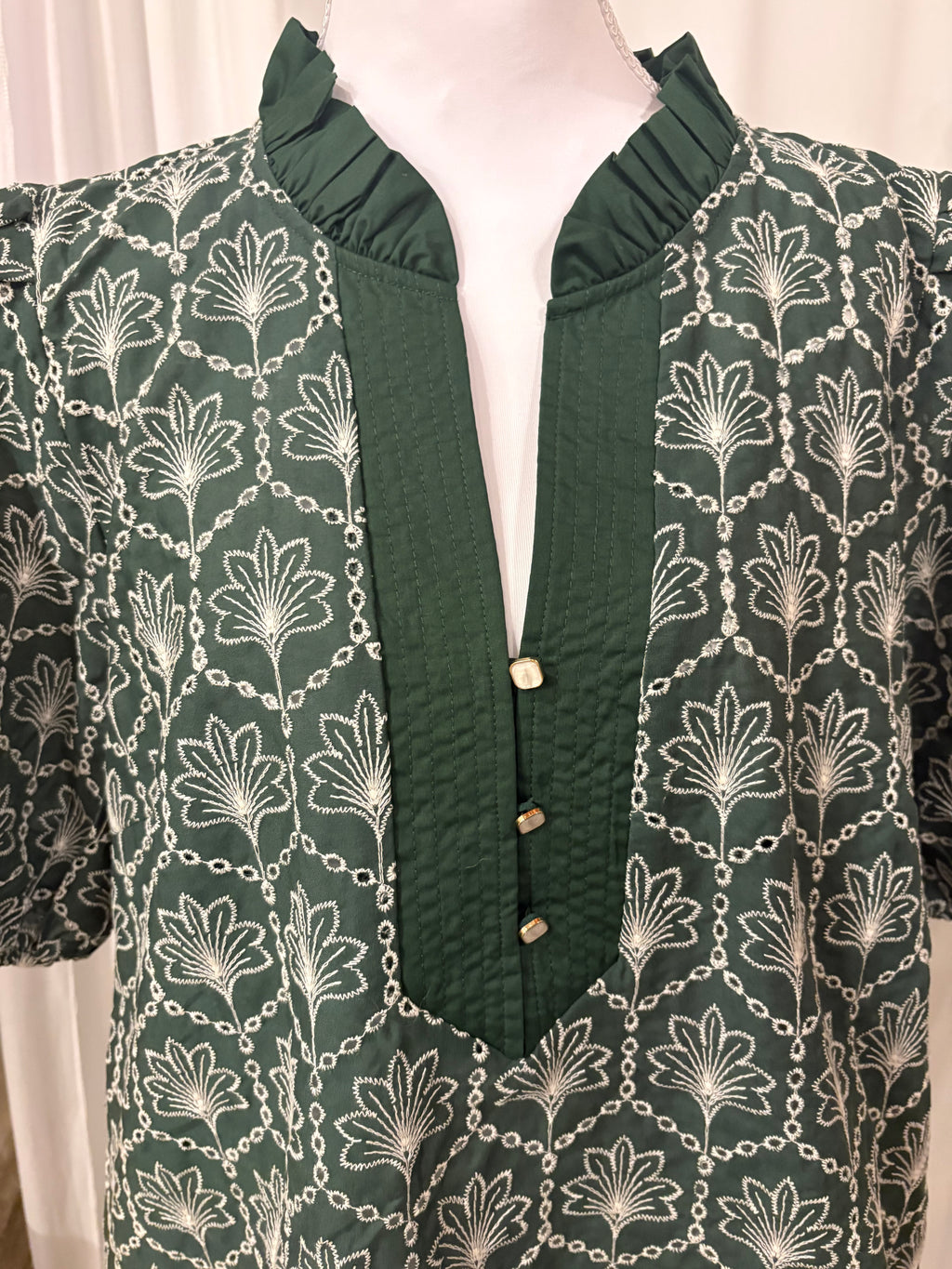 A dark green blouse with a white floral and chain pattern, featuring a ruffled mock neck and a buttoned front placket, displayed on a mannequin.