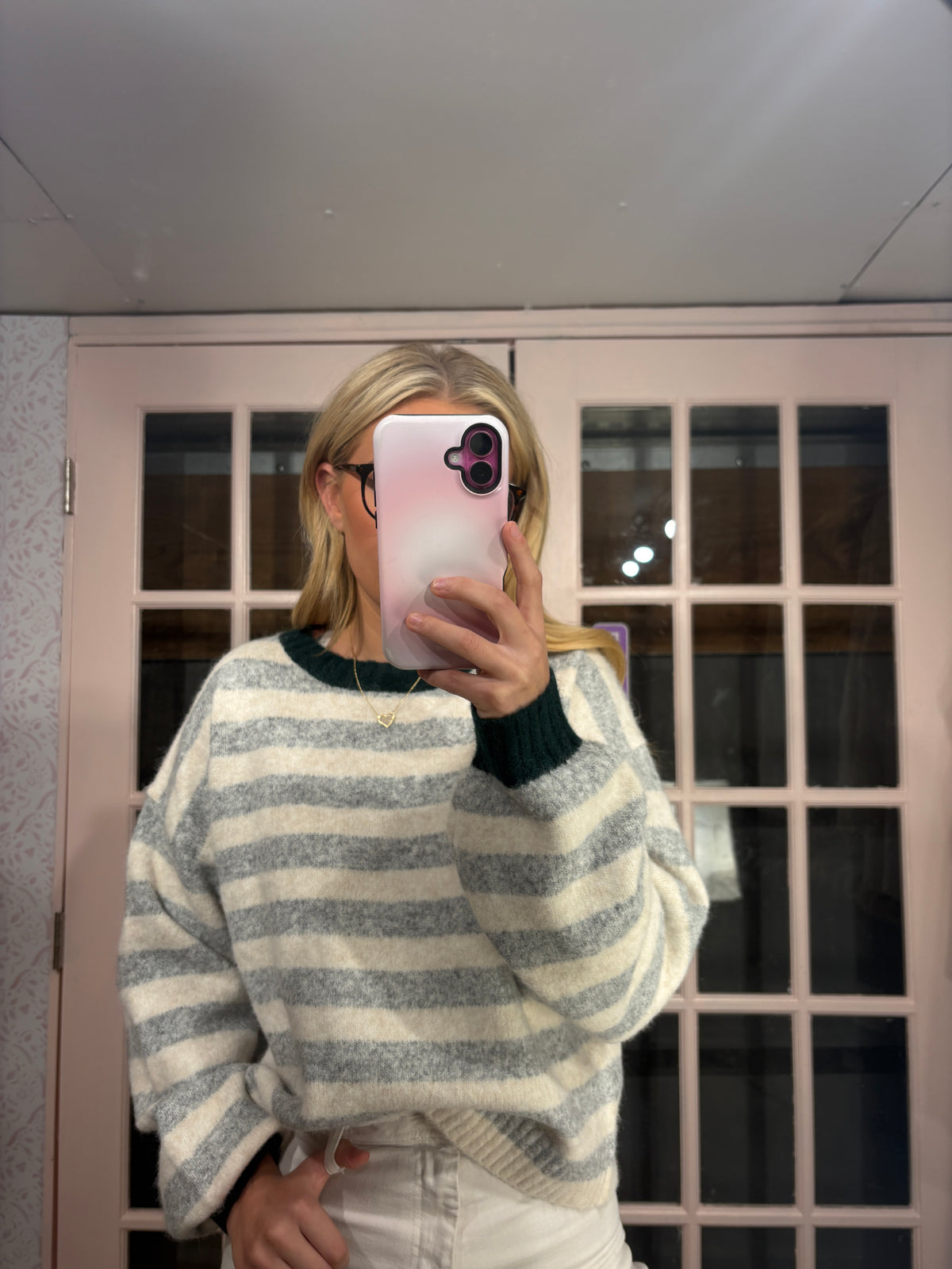 A woman takes a mirror selfie wearing a cozy, oversized knit sweater with horizontal gray and white stripes and dark green ribbed cuffs and collar. She is standing in front of a pink-framed glass door.