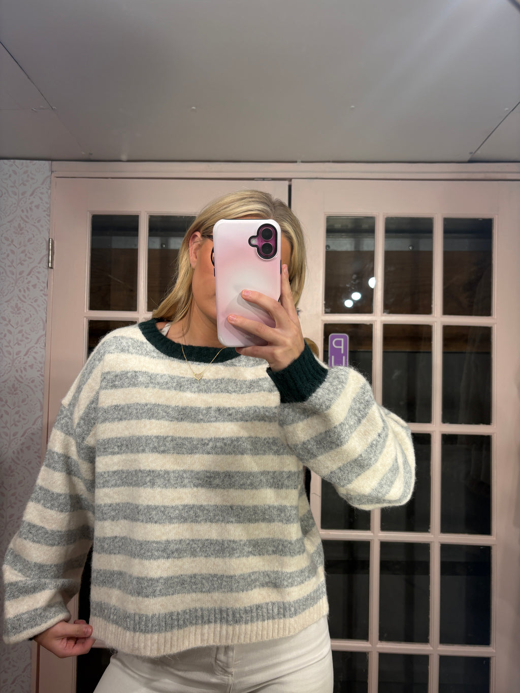 A woman takes a mirror selfie wearing a fuzzy knit sweater with horizontal gray and cream stripes and dark green ribbed trim, paired with white pants in front of a glass-paneled door.