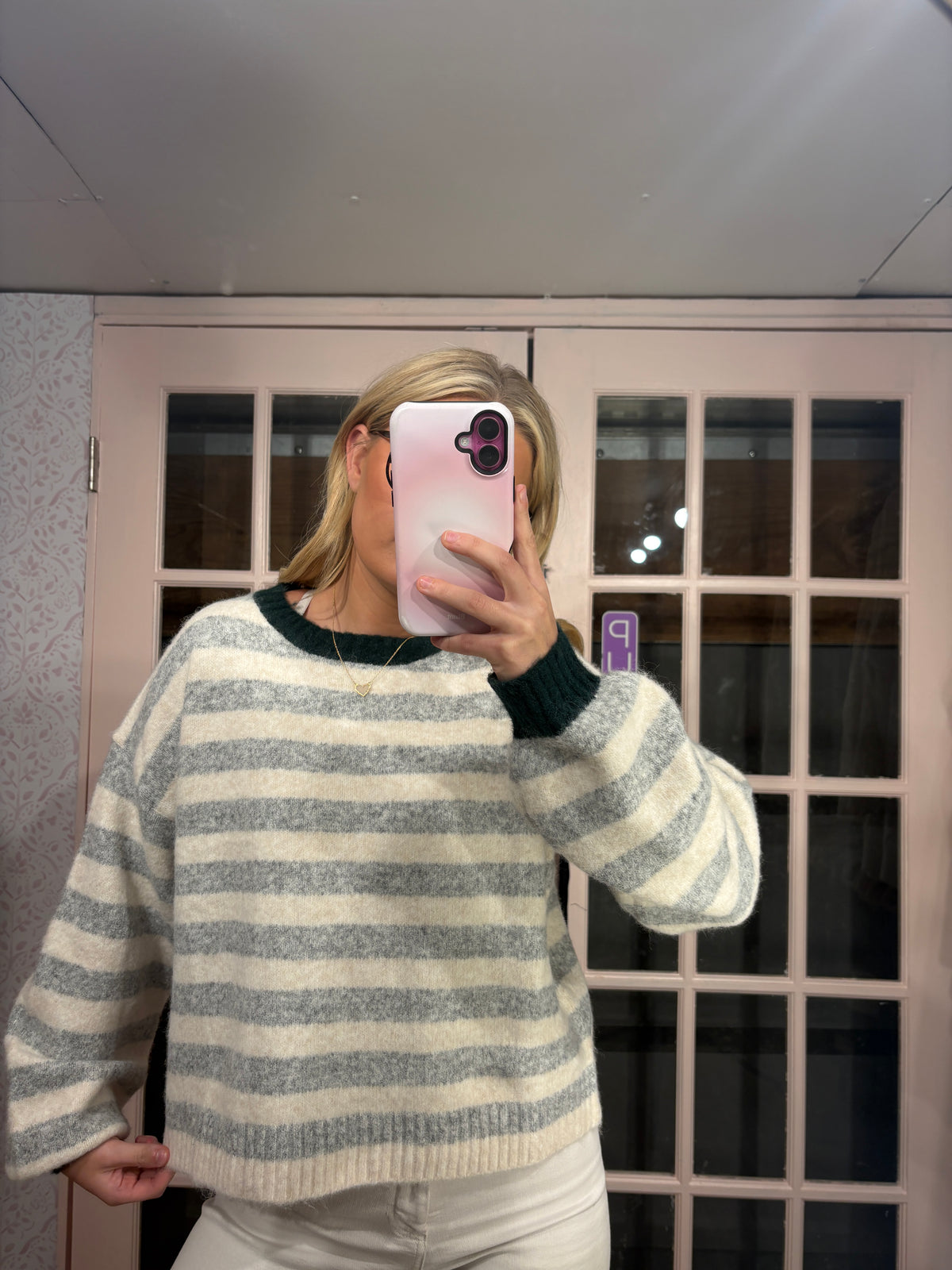 A woman takes a mirror selfie wearing a fuzzy knit sweater with horizontal gray and cream stripes and dark green ribbed trim, paired with white pants in front of a glass-paneled door.