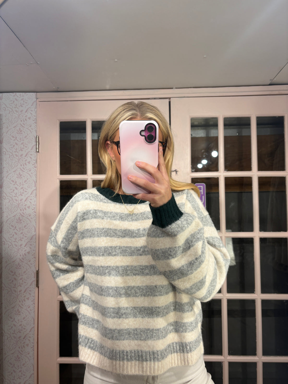 A person taking a mirror selfie wearing a fuzzy, horizontally striped knit sweater in cream and gray, with a dark green crew neck collar and cuffs. The background shows a pink-framed glass door and a light-colored ceiling.