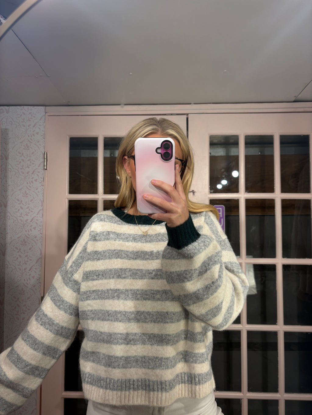 A person taking a mirror selfie wearing a gray and cream striped knit sweater with dark green ribbed collar and cuffs, holding a pink phone in front of their face.